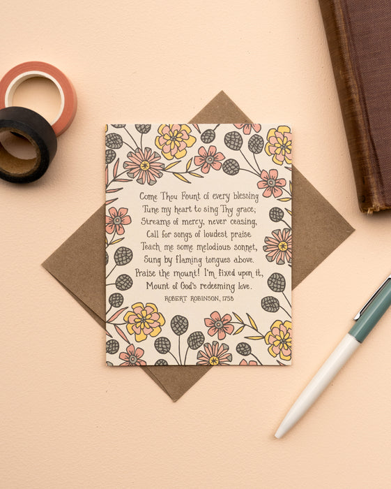 The Come Thou Fount hymn greeting card, framed by dark salmon and buttercream colored florals against a moonstone background, is styled with a kraft paper envelope, ink pen, book, and washi tape.