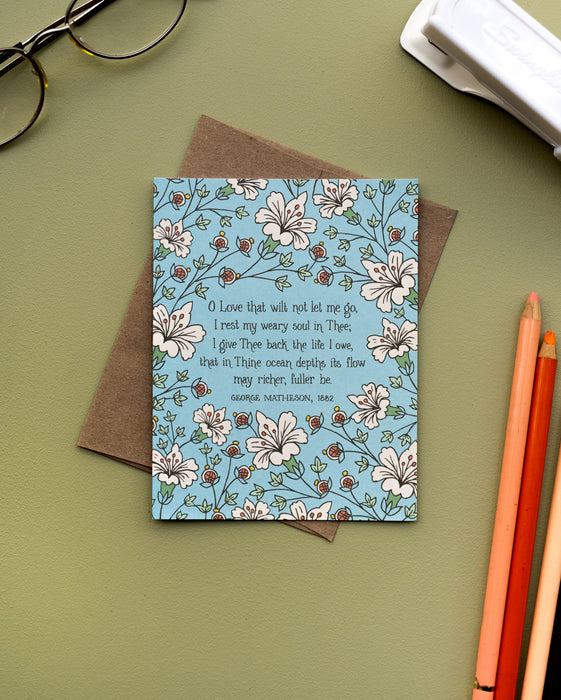 The "O Love That Wilt Not Let Me Go" hymn greeting card, framed by burnt sienna and moonstone floral against a sea mist background, styled with a recycled kraft paper envelope, colored pencils, glasses and a stapler.