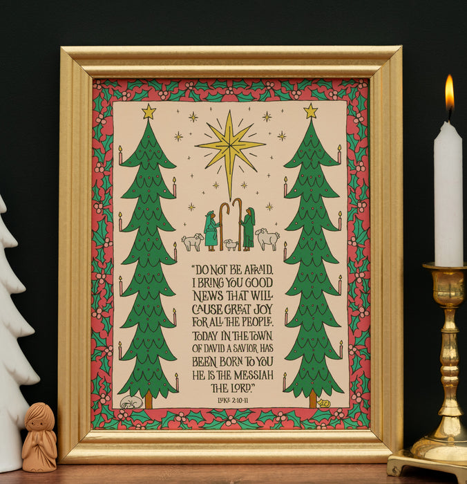 The "Good News of Great Joy" art print, which features the hand-lettered text of Luke 2:10–11 surrounded by festive trees and the wondering shepherds, is shown in a gold frame and styled with a candle and white ceramic tree and angel carving.