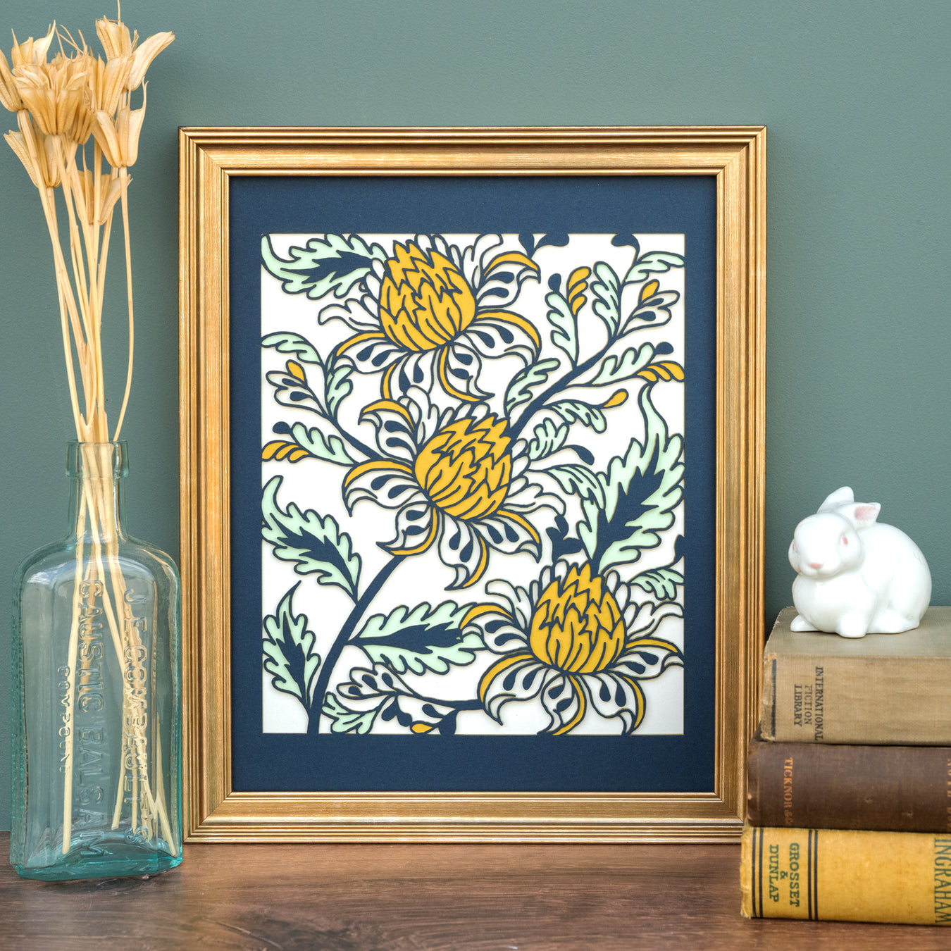 The Layered Floral Papercut “Lyra” (Spearmint) is made using cardstock in hues of flag blue, mustard, spearmint, tile green. The art is shown framed and styled with dried grasses in a vase and a stack of books with ceramic figurine.