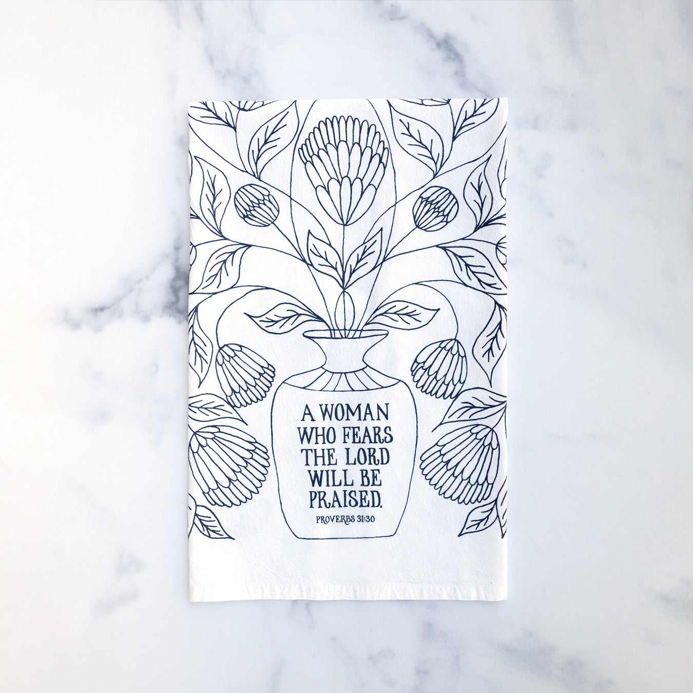 Bring wisdom and beauty into your home with The Woman Who Fears the Lord Scripture Tea Towel, featuring Proverbs 31 verses printed in midnight blue; 100% cotton. Shown folded against a marble background.