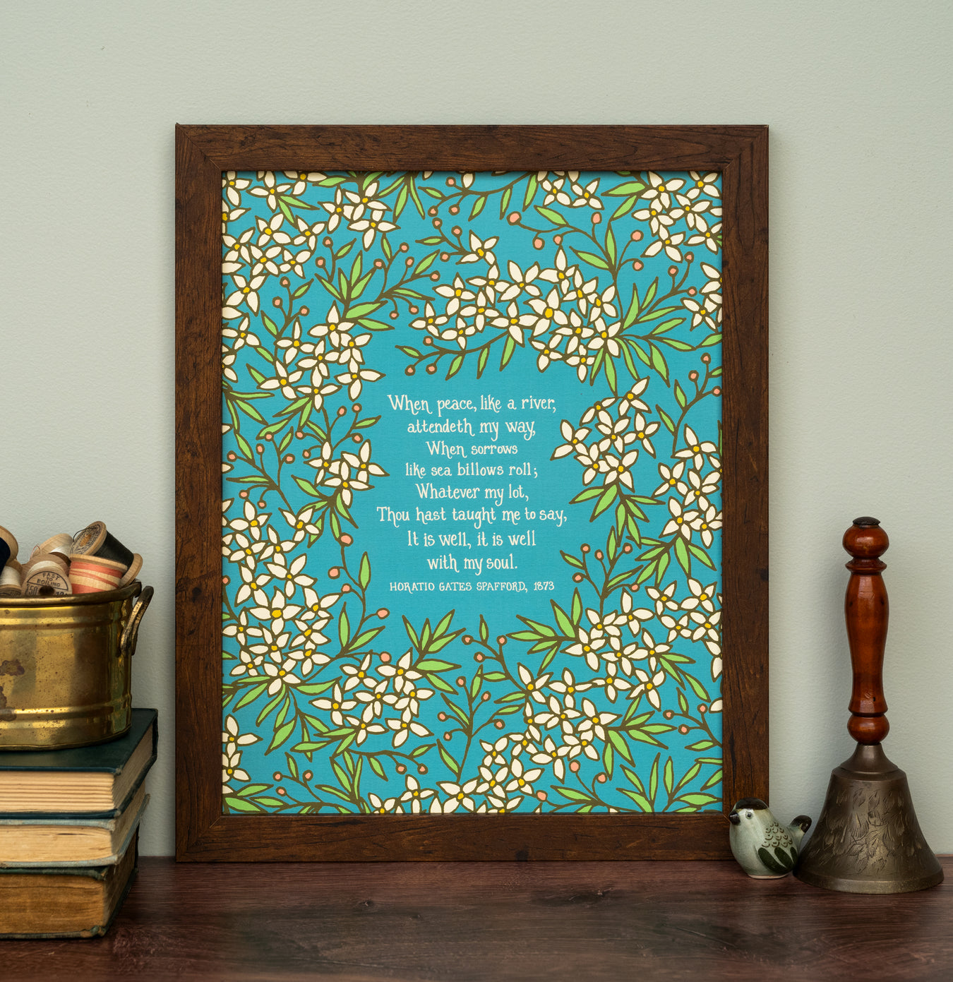 The "It Is Well With My Soul" 11" x 14" art print features a hand-lettered verse of the well-loved hymn framed by florals against a bright blue background, shown here framed and stuyled with a bell, books, and a tin of thread spools.