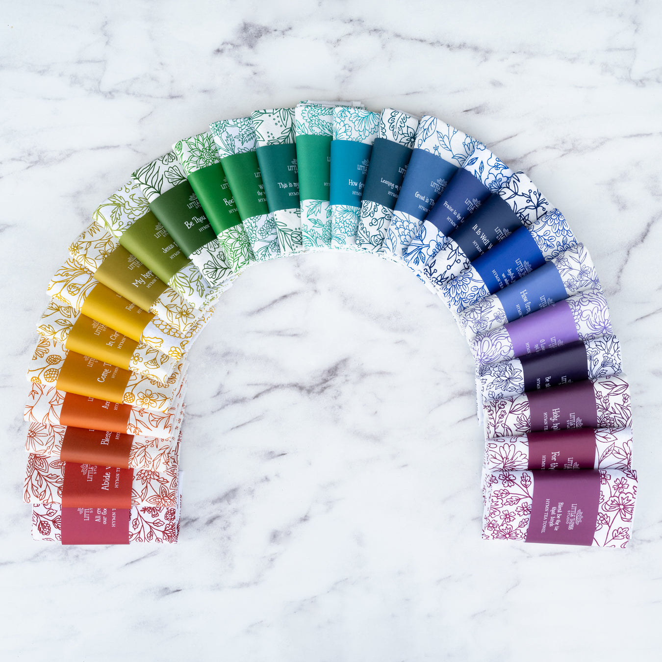 The rainbow set of 26 hymn tea towels by Little Things Studio, pictured folded and wrapped with paper belly bands for gift giving, fanned out in a rainbow of color against a white marble background.