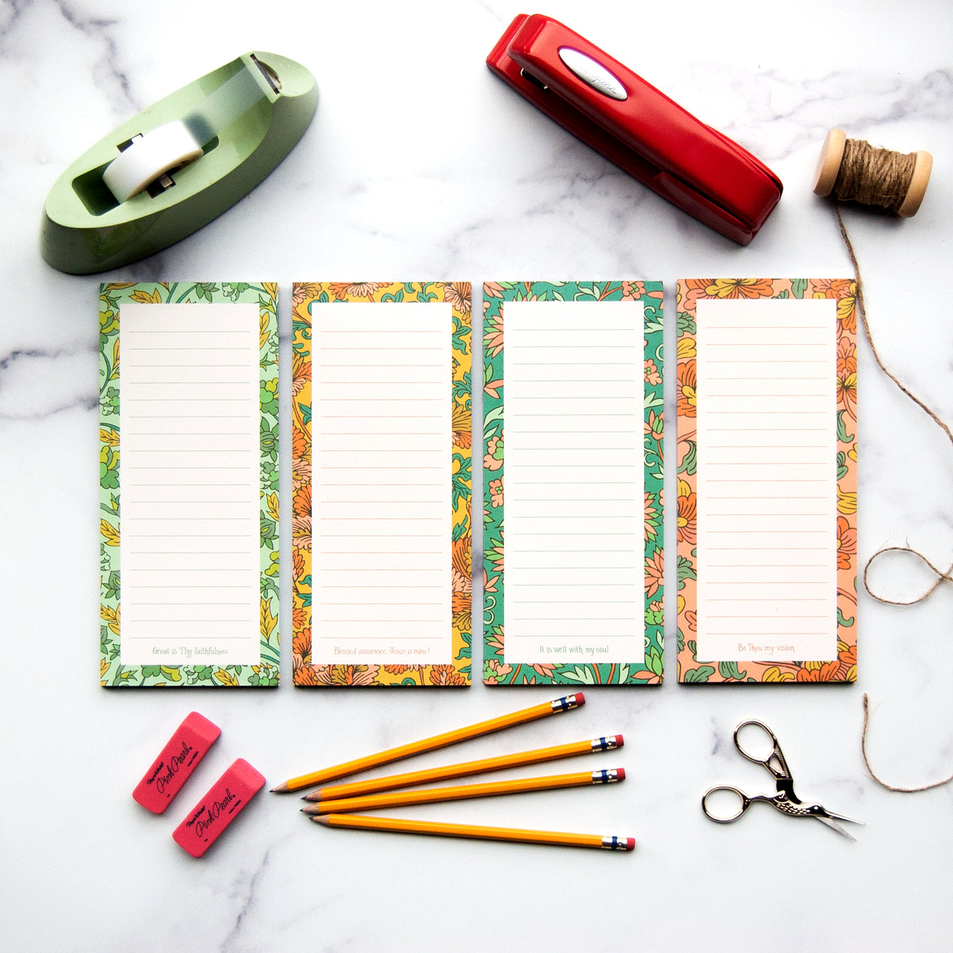 Four hymn notepads, each featuring illustrated floral borders and a line from a beloved hymn, pictured here with office supplies against a white marble background.