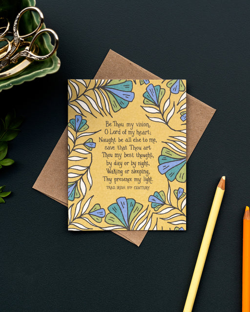 The Be Thou My Vision hymn greeting card, framed by rainforest green and stormy blue colored florals against a saffron background, is styled with a kraft paper envelope, colored pencils, and a ceramic cup with gold scissors.