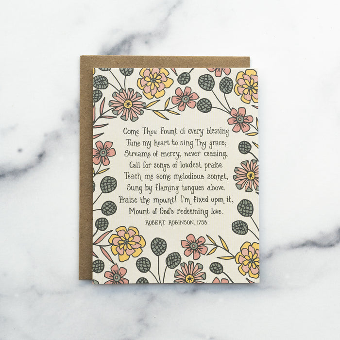 The Come Thou Fount hymn greeting card, framed by dark salmon and buttercream colored florals against a moonstone background, shown with a kraft paper envelope against a marble background.