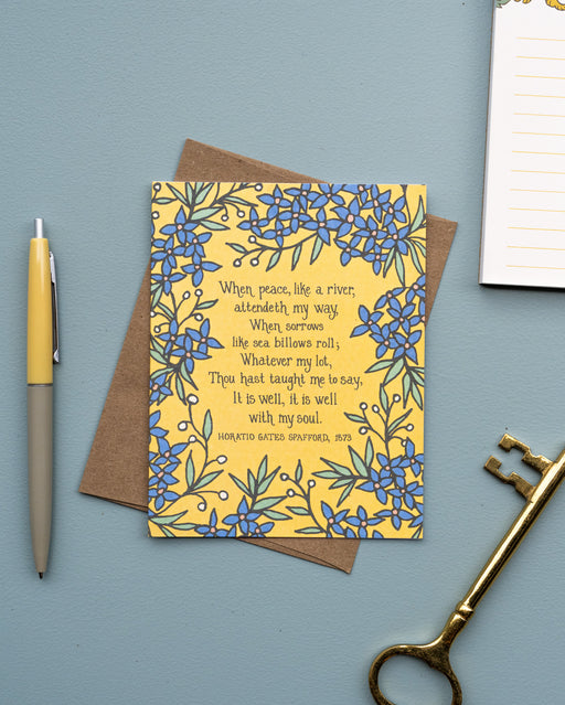 The It Is Well With My Soul hymn greeting card, framed by stormy blue and lime sorbet colored florals against a buttercream background, is styled with a kraft paper envelope, ink pen, notepad, and a gold skeleton key.