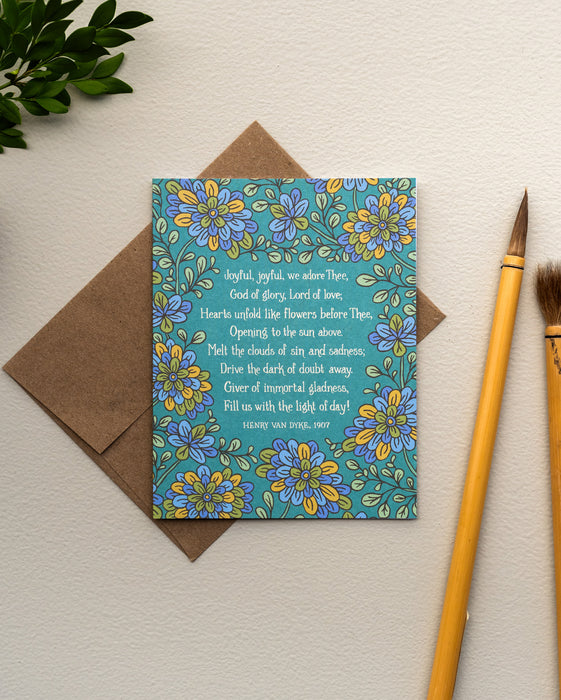 The "Joyful, Joyful, We Adore Thee" hymn greeting card, framed by chartreuse and cornflower floral against a rainforest green background, is styled with a kraft paper envelope, greens, and paintbrushes.