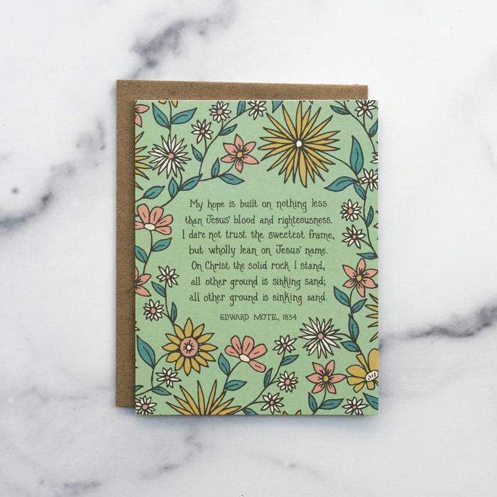 The My Hope Is Built hymn greeting card, framed by saffron and dark salmon colored florals against a lime sorbet background, shown with kraft envelope against a marble background.