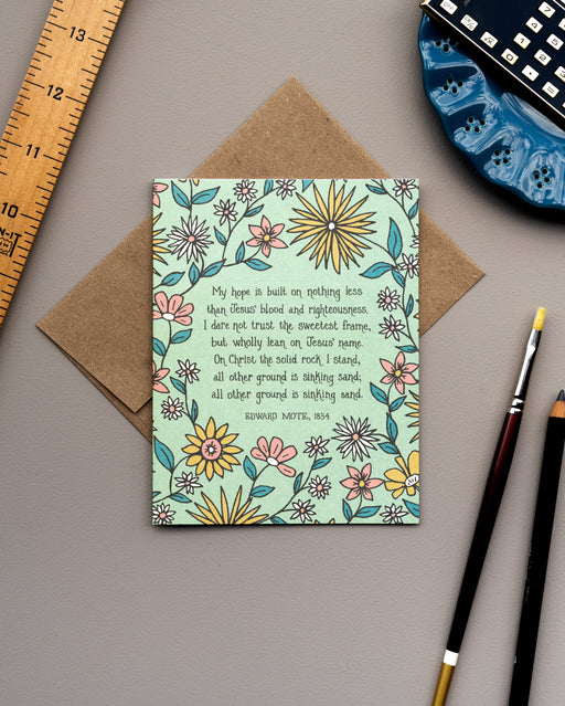 The "My Hope Is Built" hymn greeting card, framed by saffron and dark salmon colored florals against a lime sorbet background, is styled with a kraft envelope, paintbrush and pencil, and a wooden ruler.