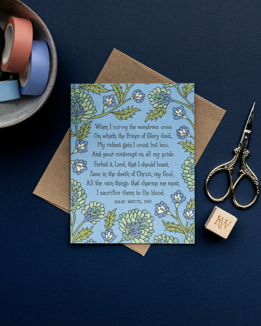 The When I Survey the Wondrous Cross hymn greeting card, framed by chartreuse and lime sorbet floral against a cornflower background, is styled with a kraft paper envelope, scissors, a stamp, and washi tape.