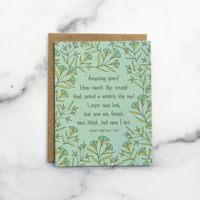 The Amazing Grace hymn greeting card, framed by dark mint and vintage yellow floral against a baby green background, shown with kraft paper envelope against a marble background.