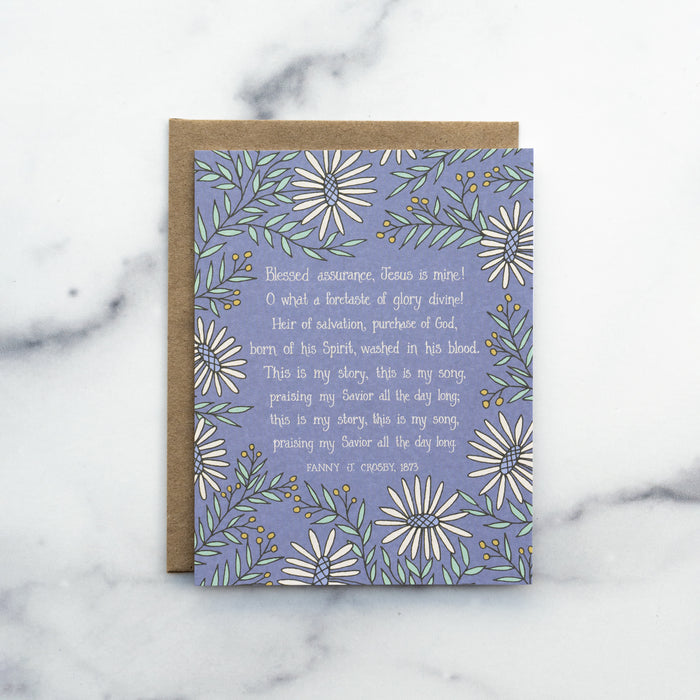 The Blessed Assurance hymn greeting card, framed by moonstone and baby green floral against a lavender gray background, shown with a kraft paper envelope against a marble background.