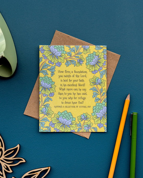 The How Firm a Foundation hymn greeting card, framed by asparagus and lilac colored florals against a vintage yellow background, is shown here styled with kraft paper envelope and pencils against a deep teal background.