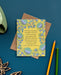 The How Firm a Foundation hymn greeting card, framed by asparagus and lilac colored florals against a vintage yellow background, is shown here styled with kraft paper envelope and pencils against a deep teal background.