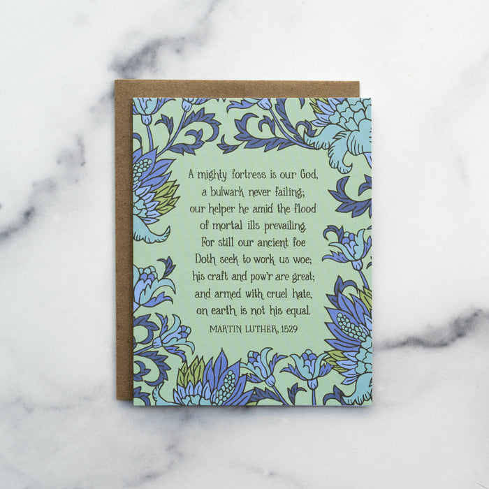 The A Mighty Fortress greeting card, framed by thunder blue and fern floral against a pastel green background, shown with a kraft paper envelope against a marble background.