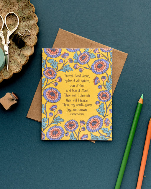 The Fairest Lord Jesus hymn greeting card, framed by blue violet and burnt sienna florals against a sunlight background, is styled with a kraft paper envelope, colored pencils, and a ceramic dish with scissors and clips.