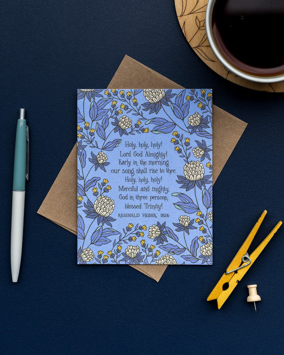 The Holy, Holy, Holy! hymn greeting card, framed by thunder blue and moonstone floral against a summer sky background, is styled with a recycled kraft paper envelope, clothes pin and thumb tack, ink pen, and a ceramic mug.