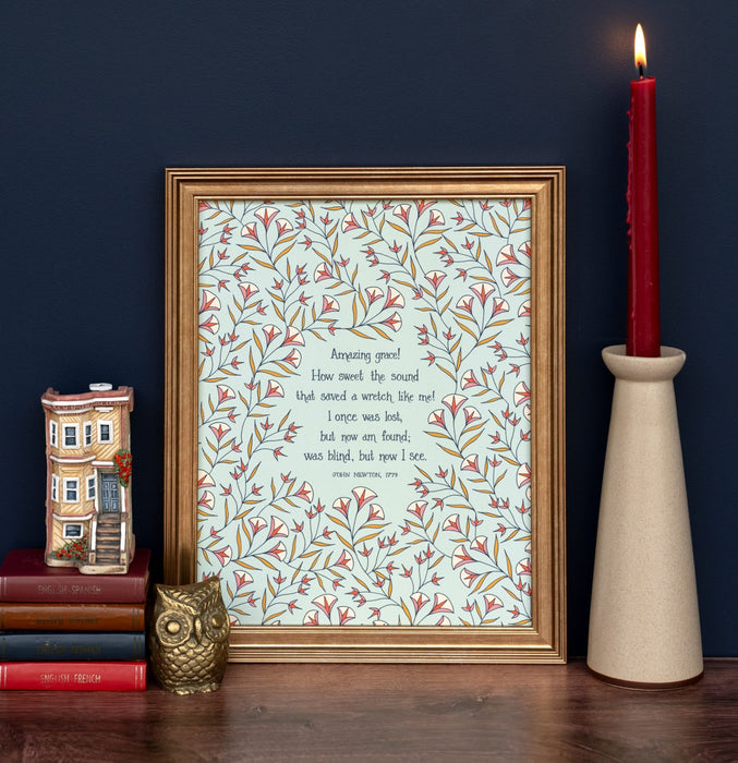 Amazing Grace lyrics wall art featuring hand lettered text on a sea foam green background surrounded by delicate illustrated florals in light red, yellow, and cream, displayed in a gold frame with a ceramic taper candle holder, stack of books and decorative ceramic figurines.