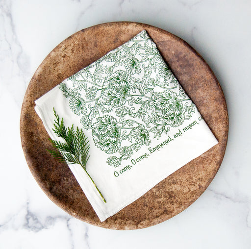 "O Come, O Come Emmanuel" cloth napkins from Little Things Studio printed in festive evergreen with a floral illustration and hymn text around the edge. Shown folded square and styled against a wooden plate with a sprig of greenery.