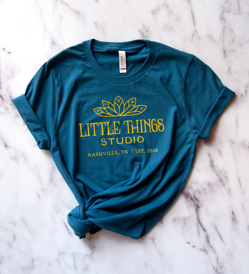 Little Things Studio t-shirt in deep teal, featuring our logo in mustard yellow. Printed on a super comfy Unisex Jersey Bella+Canvas short sleeve tee. Shown against a marble backdrop.