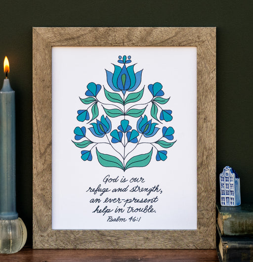 The "God Is Our Strength" Scripture art print features the hand-lettered verse Psalm 46:1 is accented by a blue and green floral motif against a cotton white background, shown in a wood frame styled with a candle, books, and a figurine.