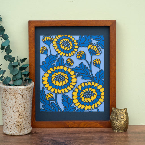 The Layered Floral Papercut “Lou” (Cornflower) is made using cardstock in hues of flag blue, Prussian blue, light gold, cornflower. Shown here framed and styled with a vase of greens and ceramic figurine.