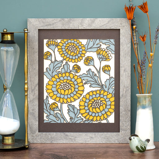 The Layered Floral Papercut “Lou” (Fudge) is made using cardstock in hues of hot fudge, steel blue, mustard, cream. Shown here styled with an hour glass, flower vase and ceramic figurine.