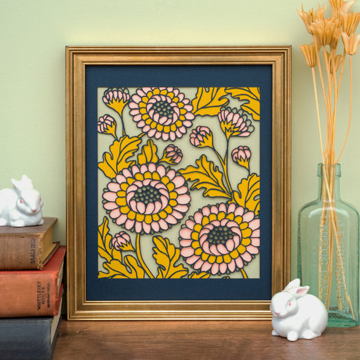 The Layered Floral Papercut Lou in Kiwi is made using cardstock in hues of flag blue, mustard, salmon, and kiwi, shown here framed and styled with a vase of dried grasses, a stack of books, and two ceramic rabbits.