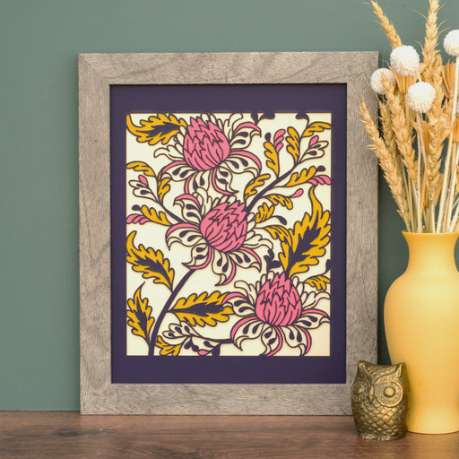 The Layered Floral Papercut “Lyra” (Violet) is made using cardstock in hues of violet, deep pretty pink, mustard, and cream, shown here framed and styled with a vase of dried flowers and a brass figurine.