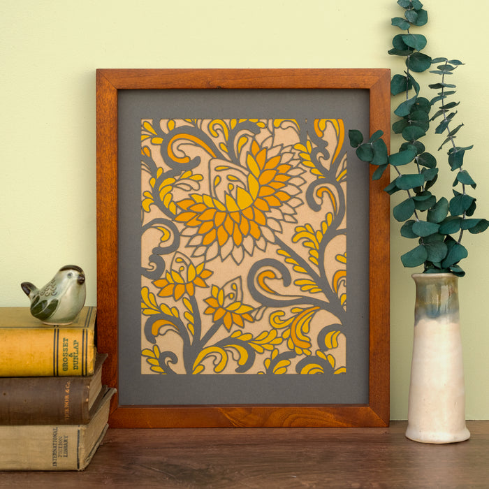 The Layered Floral Papercut “Margot” (Dark Gray) is made using cardstock in hues of dark gray, pumpkin pie, mustard, and kraft, shown framed and styled with a vase of eucalyptus and a stack of books with bird figurine.