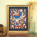 The Layered Floral Papercut “Margot” (Salmon) is made using cardstock in hues of flag blue, tangy orange, deep ocean blue, salmon, shown framed and styled with a glass vase of dried grasses, books, and a wooden figurine.