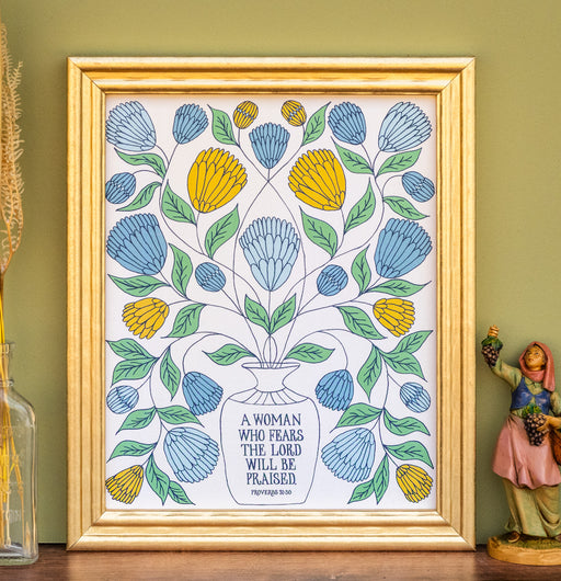 "A Woman Who Fears" Scripture art print features the hand-lettered verse accented by a spray of light blue, golden, and dusk blue florals with a cream background. Shown framed and styled with a vase of dried grasses and a ceramic figurine.