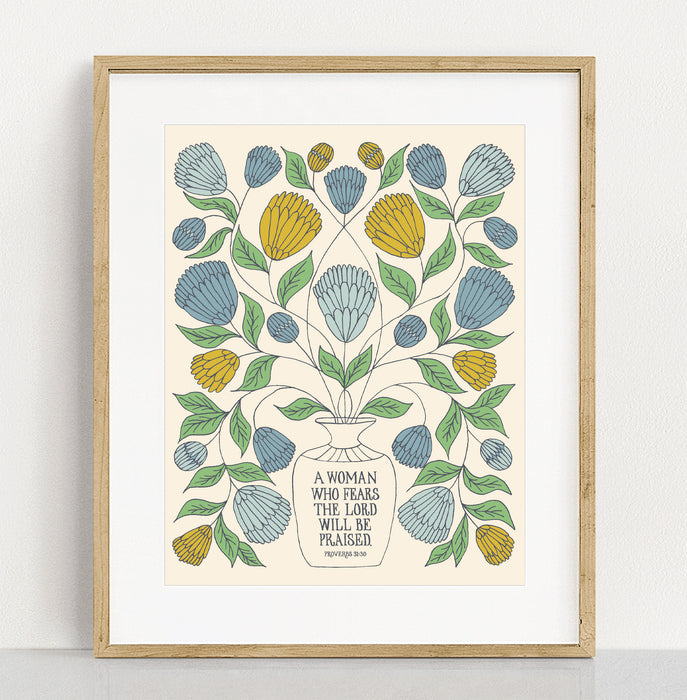 "A Woman Who Fears" Scripture art print in 11x14 features the hand-lettered verse accented by a spray of light blue, golden, and dusk blue florals with a cream background. Shown in a wooden frame against a white background.