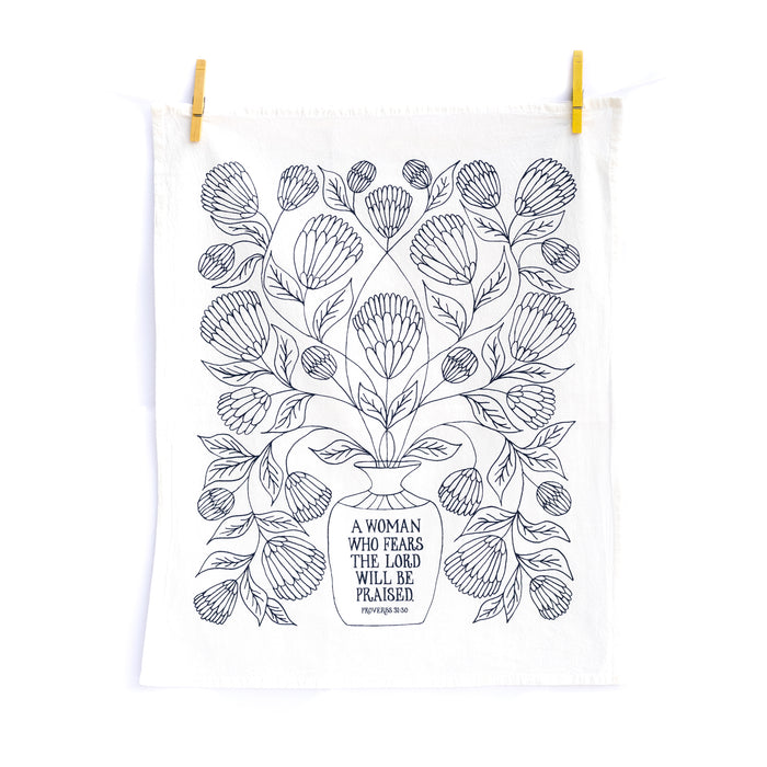 Bring wisdom and beauty into your home with The Woman Who Fears the Lord Scripture Tea Towel, featuring Proverbs 31 verses printed in midnight blue; 100% cotton. Shown unfolded and hanging with clothes pins.