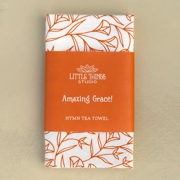 The Amazing Grace hymn tea towel is printed in pumpkin orange and features hand illustrated floral designs surrounding the hand lettered text, shown folded and wrapped with a belly band for gift giving and displayed against a khaki background.