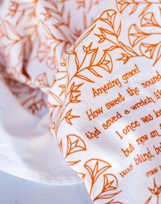 Floral and text detail of the Amazing Grace tea towel is one of our hymn tea towels, printed in pumpkin orange and featuring hand illustrated floral designs surrounding the hand lettered text