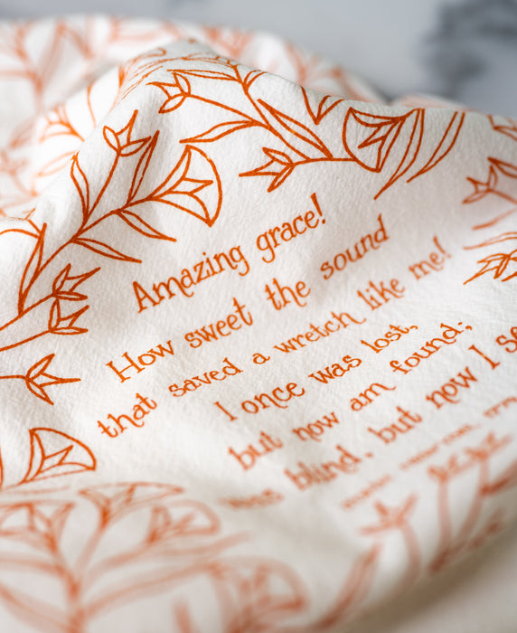 Floral and text detail of the Amazing Grace tea towel is one of our hymn tea towels, printed in marmalade orange and featuring hand illustrated floral designs surrounding the hand lettered text.