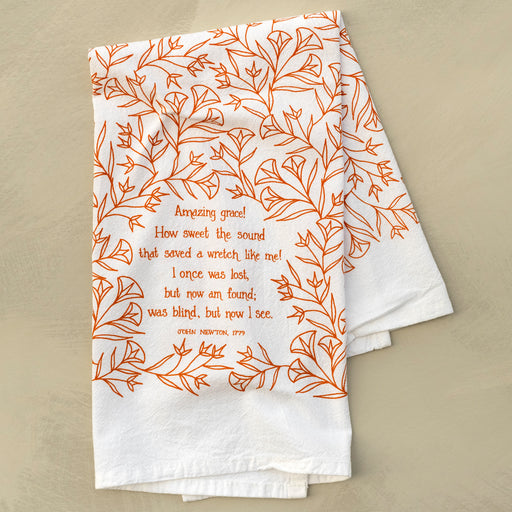 The Amazing Grace hymn tea towel is printed in pumpkin orange and features hand illustrated floral designs surrounding the hand lettered text, shown folded and displayed against a khaki background.