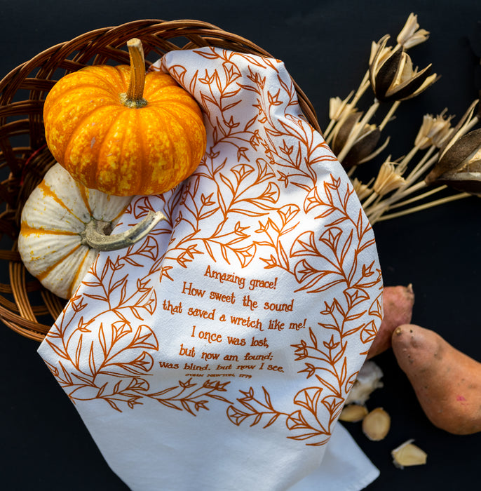 Amazing Grace tea towel is one of our hymn tea towels, printed in pumpkin orange and featuring hand illustrated floral designs surrounding the hand lettered text, shown styled with mini pumpkins and dried grasses.