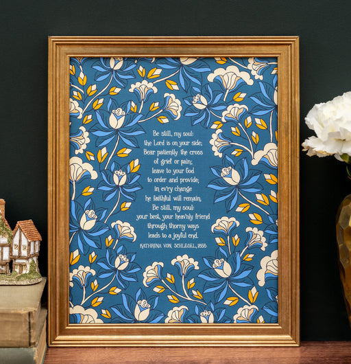 The 8x10 "Be Still, My Soul" hymn art print features hand-lettered hymn text surrounded by a detailed floral on a navy blue background. Shown in a gold frame and styled with a vase and flower, as well as a ceramic house figurine.