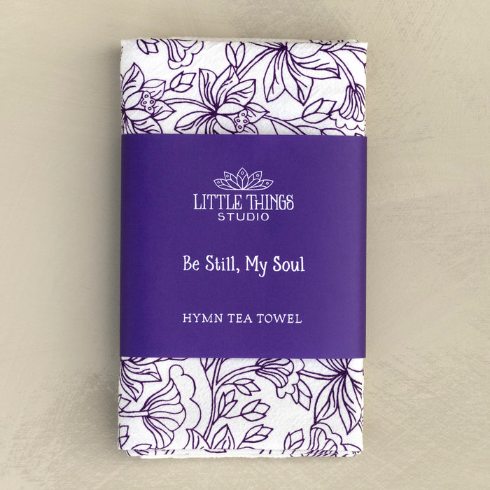 The Be Still, My Soul tea towel featuring the beloved hymn is printed in a deep eggplant purple, shown folded against a khaki background and wrapped with a paper belly band for gift giving.