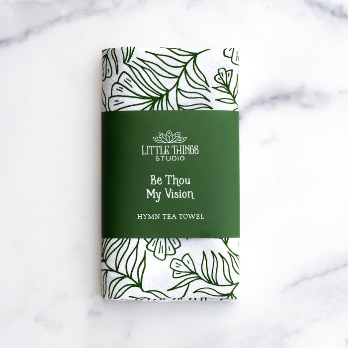 The Be Thou My Vision tea towel is printed in forest green and features hand lettered text surrounded by illustrated botanical detail, shown folded and wrapped with a paper belly band for gift giving.