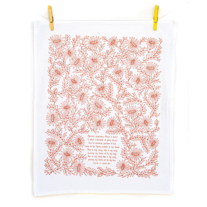 The Blessed Assurance tea towel, printed in rusty red and featuring hand lettered hymn text surrounded by intricate floral illustration, is shown unfolded and hanging with clothes pins