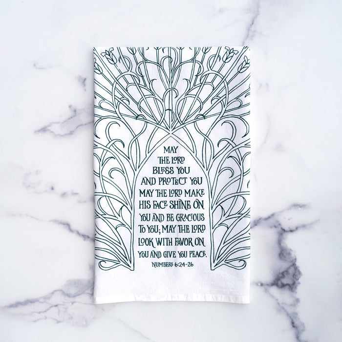 The Blessing Scripture tea towel is printed in muted blue-green tone and features the verse from Numbers 6:24–26. Made of 100% cotton. Shown here folded against a marble background.