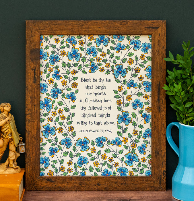 Blest be the Tie hymn art print, featuring hand lettered text offset by delicate hand illustrated floral designs, printed on a cream background, displayed in a dark wood frame with a pitcher of greens and ceramic figurine.