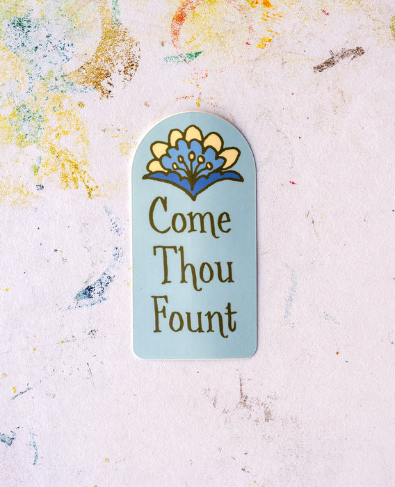 Hymn stickers from Little Things Studio are here! This Come Thou Fount hymn sticker features a light blue background and a single bloom in hues of blue and cream; displayed with a work table as the background.