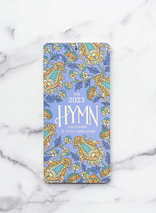 The 2023 Hymn Calendar by Little Things Studio, featuring 12 months of hymns with hand lettered text and vibrant floral illustration, displayed here showing the cover against a white marble backdrop.