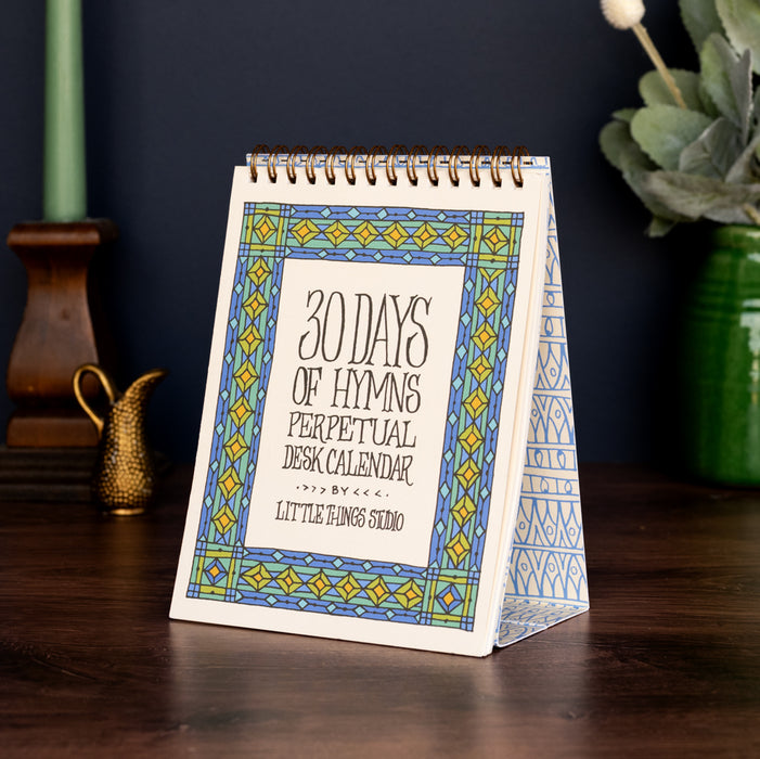 A refreshed 30 days of hymns perpetual calendar on an easel with intricate illustrations to accent hand-lettered hymn texts for each day, shown styled with a lit candle, vase wtih greens, and a small pitcher.