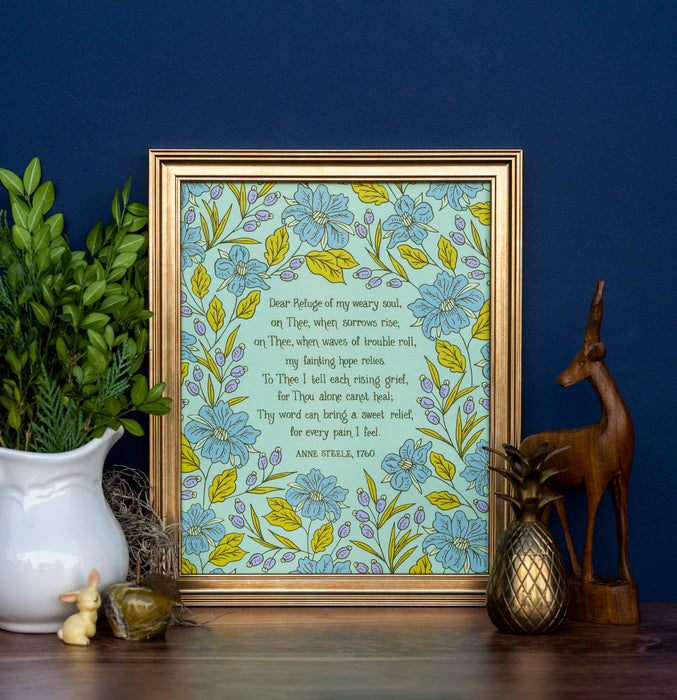 The "Dear Refuge Of My Weary Soul" hymn art print, surrounded by a gorgeous blue and lavender floral with a light teal background, is displayed in a gold frame alongside a vase of fresh greens and various figurines, against a navy blue wall.